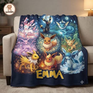 May include: A dark blue fleece blanket featuring a colorful graphic print of eight different Eevee evolution characters arranged around a central Eevee. The name EMMA is printed in large yellow block letters at the bottom of the blanket.
