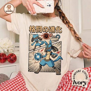 May include: An ivory t-shirt featuring a graphic print of two blue and black canine-like creatures. The text above the graphic reads "格闘の進化". One creature is smaller, while the larger one holds a glowing blue sphere. The design includes a fist icon and gear motifs.