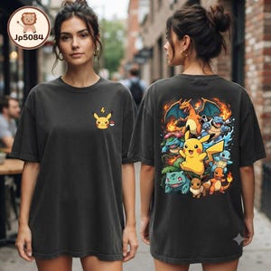 May include: A charcoal gray t-shirt featuring a small Pikachu graphic on the front left chest and a large, colorful illustration on the back displaying multiple characters including Pikachu, Charizard, Blastoise, Venusaur, and Eevee.