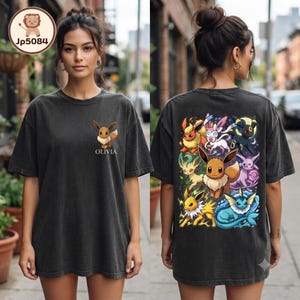 May include: A charcoal gray t-shirt featuring a small Eevee graphic and the name Olivia on the front left chest. The back displays a large, colorful illustration of Eevee and its various evolutions, including Flareon, Sylveon, and Umbreon.