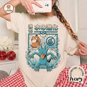 May include: An ivory t-shirt featuring a graphic print of a brown duck-like creature and a blue water-type creature with Japanese text above them. The design includes a water droplet icon and a circular sphere, all framed in a rectangular border.
