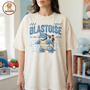 May include: A cream-colored short-sleeve t-shirt featuring a graphic of the character Blastoise. The design includes the text "BLASTOISE" and "EST. 1996" in blue, accompanied by a checkered pattern and two faint outline sketches of the same character.