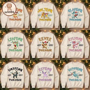 May include: A collection of nine cream-colored crewneck sweatshirts, each featuring a different Pokemon character with a holiday theme, the text 'est 1997', and the word 'Pokemon' printed below, displayed in a grid against a pine branch background.