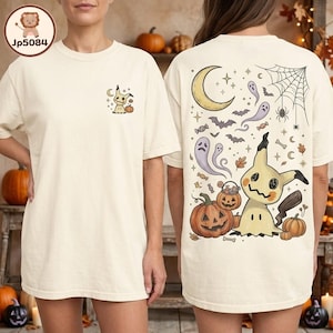 May include: An off-white t-shirt featuring a small Mimikyu and pumpkin graphic on the front left chest. The back displays a large Halloween-themed illustration with a crescent moon, ghosts, bats, a spider web, candy, and a large Mimikyu.