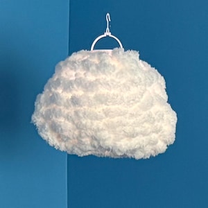 Könnte beinhalten: Eine wolkenförmige Pendelleuchte. Das Licht ist von einem flauschigen, weißen Material umgeben, das einer Wolke ähnelt. Die Leuchte hängt an einem weißen Metallhaken vor einer blauen Wand.