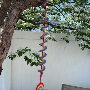 May include: A long, hanging crochet decoration featuring a spiral design in rainbow colors including red, orange, yellow, green, blue, and purple. The spiral structure is suspended vertically and terminates in a small, semi-circular rainbow motif.