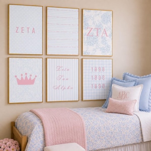 May include: Six framed wall art prints with light blue and pink designs featuring the text ZETA, ZTA, Zeta Tau Alpha, and 1898, alongside a pink crown graphic. The prints are displayed in a grid above a bed with blue floral bedding and pink accents.