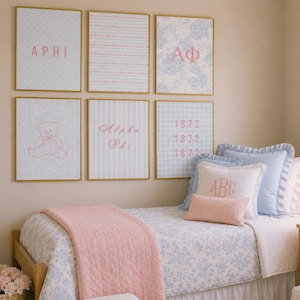 Op de afbeelding: Een slaapkamer met een eenpersoonsbed met een lichtblauwe bloemendeken, een roze doorgestikte sprei en diverse kussens, waaronder een met het monogram ABC. Zes ingelijste prints aan de muur tonen de tekst APHI, Alpha Phi en 1872.