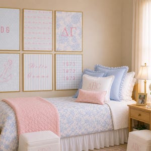 May include: A twin bed with a blue floral quilt, a pink quilted throw, and several pillows, including one with the letters ABC. Above the bed are six framed prints featuring pink text and graphics, including DG, Delta Gamma, and 1873.