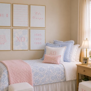 Könnte beinhalten: Ein Schlafzimmer mit einem Bett, das eine hellblaue Blumensteppdecke, eine rosa Steppdecke und mehrere Kissen aufweist. Sechs gerahmte Drucke hängen an der Wand und zeigen den Text CHI O, Chi Omega, XΩ und 1895 sowie eine Eulenillustration.