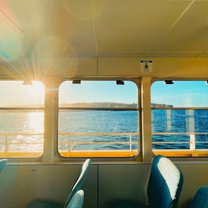 Può includere: Vista interna di un traghetto con sedili blu e finestre che si affacciano sull'oceano. Il sole splende attraverso le finestre, creando un bagliore caldo e dorato. L'acqua è blu scuro e il cielo è limpido.