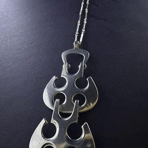 May include: A silver-toned metal pendant necklace featuring two stacked, openwork geometric shapes with circular cutouts. The pendant is suspended from a delicate, thin silver-toned chain with small, evenly spaced cylindrical metal accents.