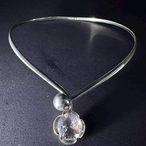 May include: A silver-toned metal collar necklace featuring a smooth, rigid band that curves into a circular shape. A small, polished silver sphere sits above a clear, faceted, flower-shaped crystal pendant that hangs from the centre of the necklace.
