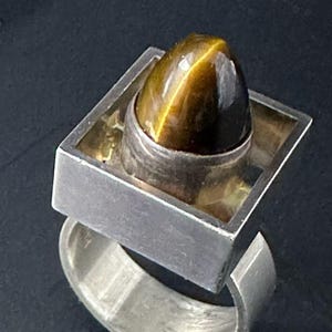 May include: A silver ring featuring a large, oval tiger's eye gemstone. The stone is set in a silver bezel, and the ring has a square design around the stone. The band is a simple, curved design.