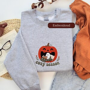 May include: A heather grey crewneck sweatshirt featuring an embroidered design of a large orange pumpkin with a smiling face. Inside the pumpkin, a small white ghost sits under a blanket reading a book. The text "cozy season" is embroidered below.