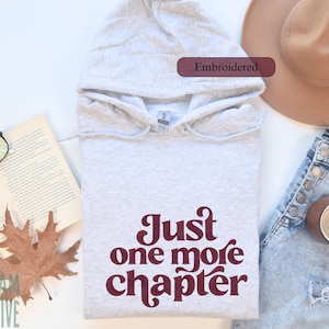May include: A light grey embroidered hoodie with the text "Just one more chapter" in burgundy. The image includes a pair of glasses, an open book, a brown hat, a pair of jeans, a cup of coffee, and a fall leaf.
