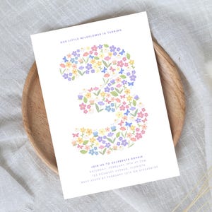 Może przedstawiać: Białe zaproszenie z tekstem "OUR LITTLE WILDFLOWER IS TURNING" i "JOIN US TO CELEBRATE SOPHIE". Liczba "3" wypełniona jest kolorowymi kwiatami. Karta leży na drewnianym talerzu.
