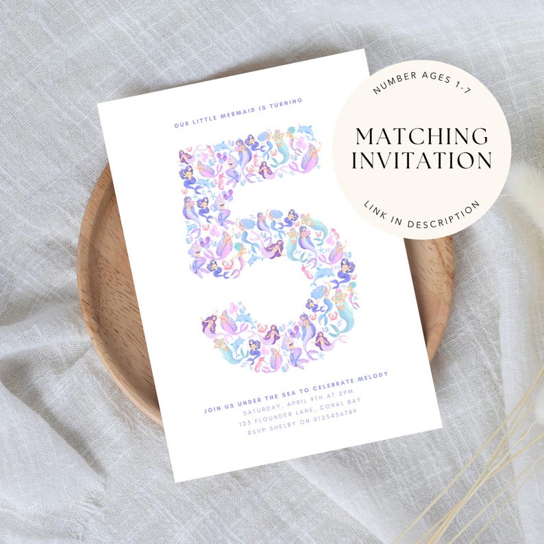 May include: A white invitation card featuring the number 5 composed of colourful mermaid illustrations. Text includes party details and "Matching Invitation". The card rests on a wooden plate. Measurements are not applicable.