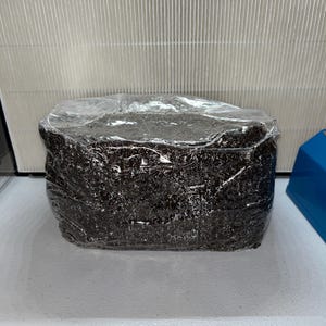 May include: A rectangular package wrapped in clear plastic. The contents appear to be dark brown and granular. The package is on a white surface, with a white air filter in the background.