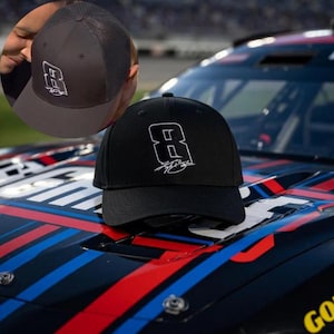 May include: Black baseball cap with the number 8 and a signature in white. The cap is displayed on a car with a black, red, and blue design. A second cap is shown in the background.