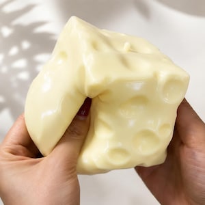 May include: A pale yellow, block-shaped object with a smooth, glossy surface and multiple circular indentations of varying sizes across its exterior. The material appears pliable and soft, showing slight deformation under pressure.