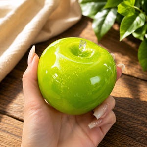 May include: A smooth, glossy, lime green glass apple sculpture featuring a clear, transparent glass stem at the top. The surface of the apple has a reflective finish with subtle internal bubbles and light-catching highlights throughout the round form.