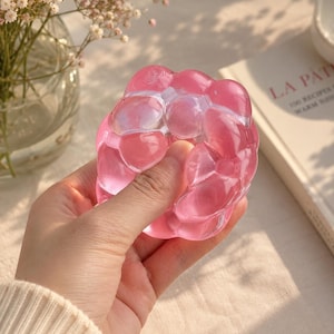 May include: A pink, translucent stress ball with a bubbly, organic shape. The stress ball is held in a hand. The background includes a vase of flowers and a book.