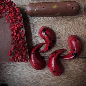 Könnte beinhalten: Eine Auswahl an handgemachten Pralinen auf einem Holzbrett. Das Bild zeigt eine Schokoladentafel mit getrockneten Himbeeren, längliche Pralinen mit Pistazien und drei rote, tropfenförmige Pralinen mit schwarzen Sprenkeln.