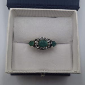 May include: An oval emerald ring with two smaller emeralds either side, surrounded by small clear stones. The ring is set in a silver band and is displayed in a white jewellery box with a blue lid.