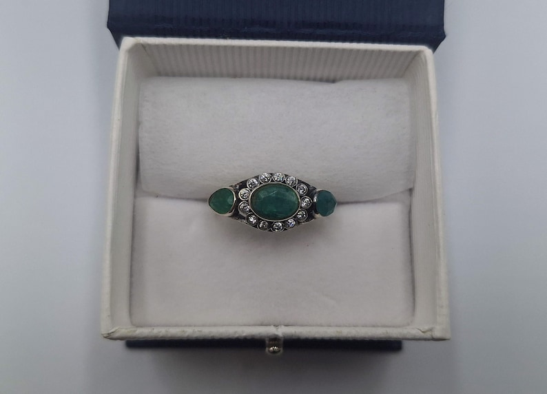 May include: An oval emerald ring with two smaller emeralds either side, surrounded by small clear stones. The ring is set in a silver band and is displayed in a white jewellery box with a blue lid.
