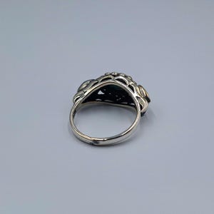 May include: A silver ring with an elaborate floral design. The ring has a dark green stone at its centre, encircled by detailed metalwork. The band is plain, polished silver.