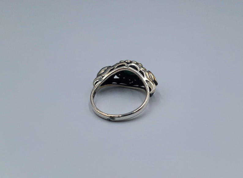 May include: A silver ring with an elaborate floral design. The ring has a dark green stone at its centre, encircled by detailed metalwork. The band is plain, polished silver.