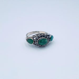 May include: A silver ring featuring three emerald-green gemstones. The central oval stone is encircled by small, clear gems. Two smaller, teardrop-shaped emeralds are either side. The ring has a detailed, openwork design.