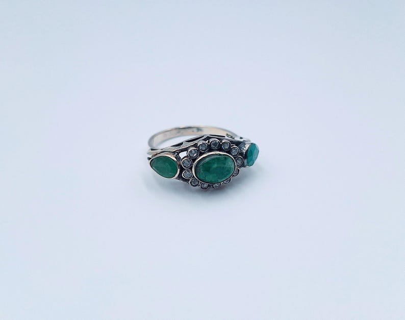 May include: A silver ring featuring three emerald-green gemstones. The central oval stone is encircled by small, clear gems. Two smaller, teardrop-shaped emeralds are either side. The ring has a detailed, openwork design.