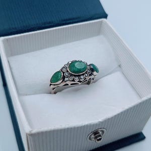 May include: A silver ring featuring three emerald-cut green gemstones and small clear stones, set in a decorative pattern. The ring is presented in a white box with a blue lid, against a light blue backdrop.