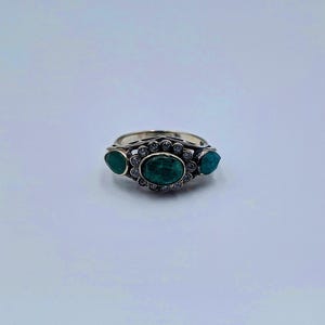 May include: A silver ring with three emerald-green gemstones. The central oval stone is encircled by small, clear accent stones. Two smaller, teardrop-shaped stones are either side of the central stone. The ring has an ornate design.