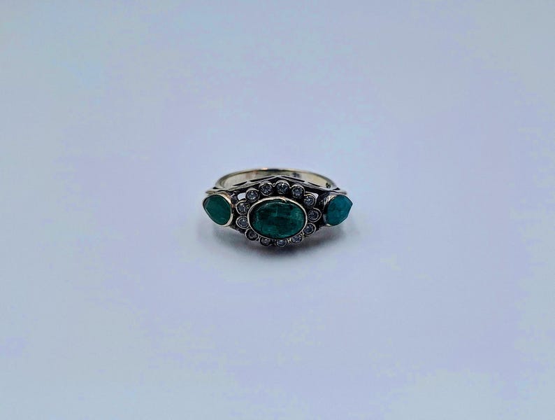 May include: A silver ring with three emerald-green gemstones. The central oval stone is encircled by small, clear accent stones. Two smaller, teardrop-shaped stones are either side of the central stone. The ring has an ornate design.