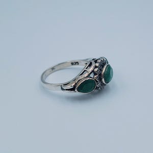 May include: A silver ring featuring three emerald-green gemstones. The ring has a decorative filigree design around the gemstones and a smooth, polished band. The ring is hallmarked with "925" on the inside of the band.