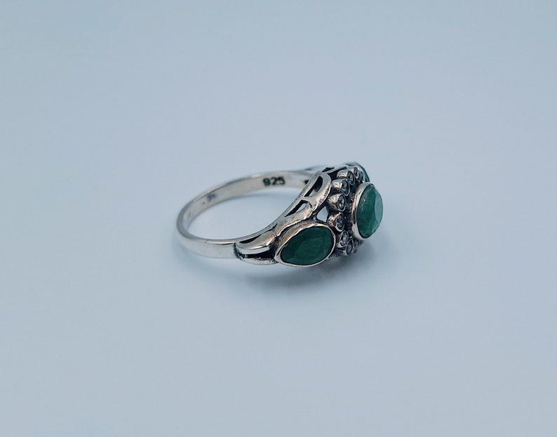 May include: A silver ring featuring three emerald-green gemstones. The ring has a decorative filigree design around the gemstones and a smooth, polished band. The ring is hallmarked with "925" on the inside of the band.