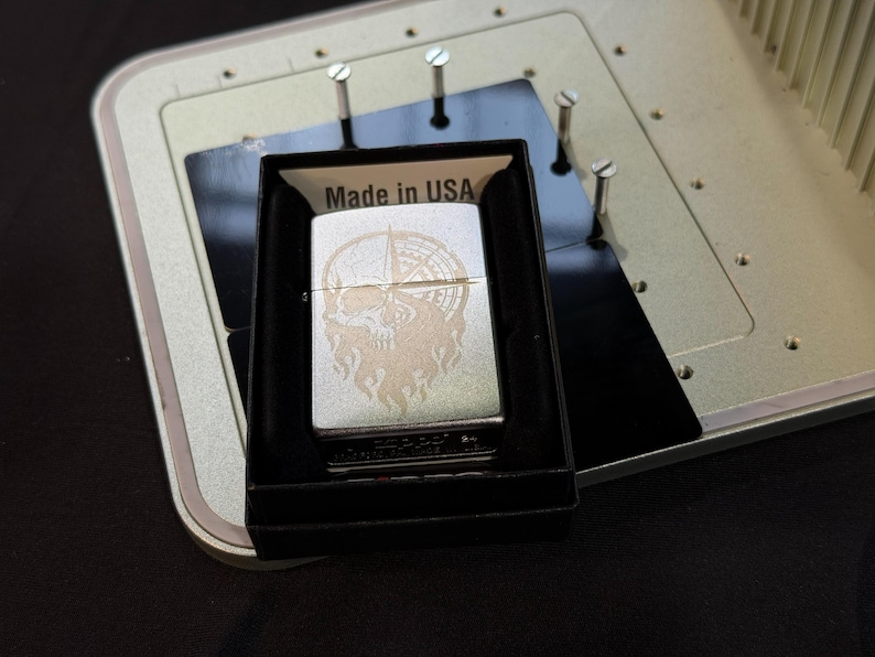 Puede incluir: Un encendedor plateado con un dise&ntilde;o de calavera y br&uacute;jula, presentado en una caja negra. El encendedor tiene un grabado detallado y el texto "Made in USA" es visible en la caja.