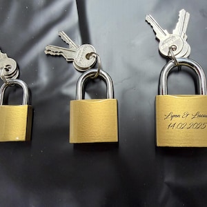 Può includere: Tre lucchetti color oro con chiavi d'argento attaccate. Il lucchetto a destra ha incisi i nomi "Lynn & Lucas" e la data "14.02.2025". I lucchetti sono su una superficie nera.