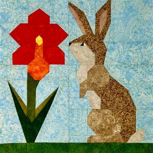 Puede incluir: Un diseño de patchwork de tela con una flor roja con un centro amarillo y un conejo marrón y canela. El fondo es azul claro con un estampado floral y la base es verde. El bloque de edredón tiene un tema de primavera o Pascua.