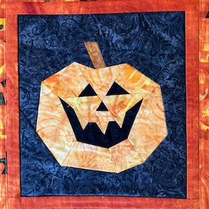 Könnte beinhalten: Ein gestepptes Wandbehang zum Thema Halloween mit einem geometrischen Kürbis-Design. Der Kürbis ist orange mit schwarzen Gesichtszügen, vor einem dunkelblauen Hintergrund. Der Rand ist orange mit Kürbismustern.