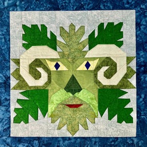 Pode incluir: Um bloco de colcha quadrado com um desenho de Green Man. O rosto é composto por várias tonalidades de tecido verde, com chifres e sobrancelhas creme. Os olhos são losangos azuis e a boca é uma meia-lua vermelha. O fundo é cinza claro com uma borda azul.