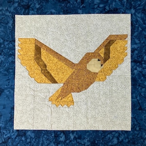 Puede incluir: Un tapiz de tela cuadrado con un diseño de acolchado de un búho de color marrón dorado en vuelo. El búho está centrado sobre un fondo gris claro moteado, enmarcado por un borde azul oscuro con un patrón texturizado en toda la tela.