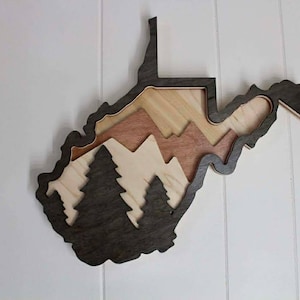 May include: Wooden wall art shaped like the state of West Virginia. The layered design features a dark gray outline, light wood, and a mountain and tree silhouette. The art piece is mounted on a white wall.
