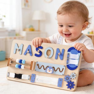 May include: A wooden activity board with the name "MASON" in blue letters. The board features interactive elements like beads, a zipper, gears, and a penguin design. Designed for early childhood development, it's a learning toy.