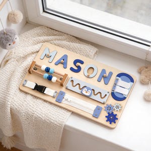 May include: Wooden activity board with the name "MASON" in blue letters. Features include a zipper, buckle, abacus, and gears. A light blue section has a penguin graphic and the word "wow".
