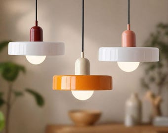 May include: Three contemporary pendant lights with a minimalist design. Each light has a coloured top, a white disc-shaped shade, and a spherical light bulb. The colours include red, orange, and peach. The lights hang from black cords.