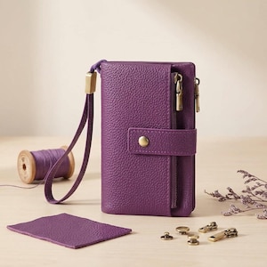 May include: A purple pebbled leather wallet with a wrist strap, a vertical zippered pocket, and a snap button closure. The wallet is accompanied by a matching purple thread spool, a small leather swatch, and several brass-toned metal hardware pieces.